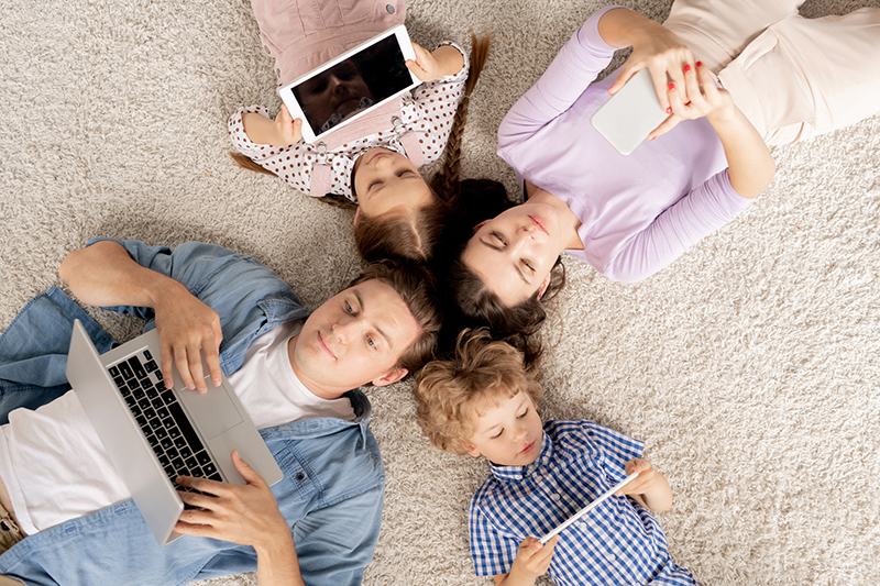 Two parents and two kids laying on the ground, holding iPads, phones and a laptop